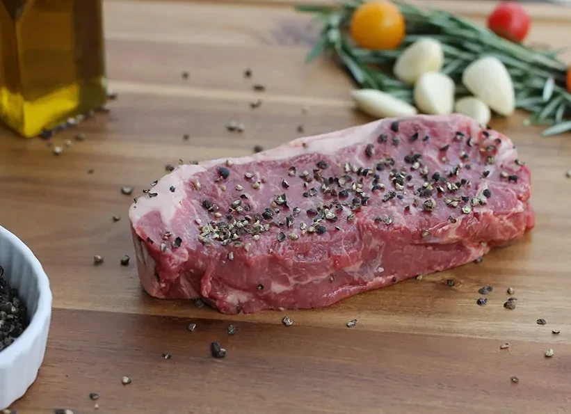 Thick raw steak, peppered, on wood with oil, garlic, and tomatoes.