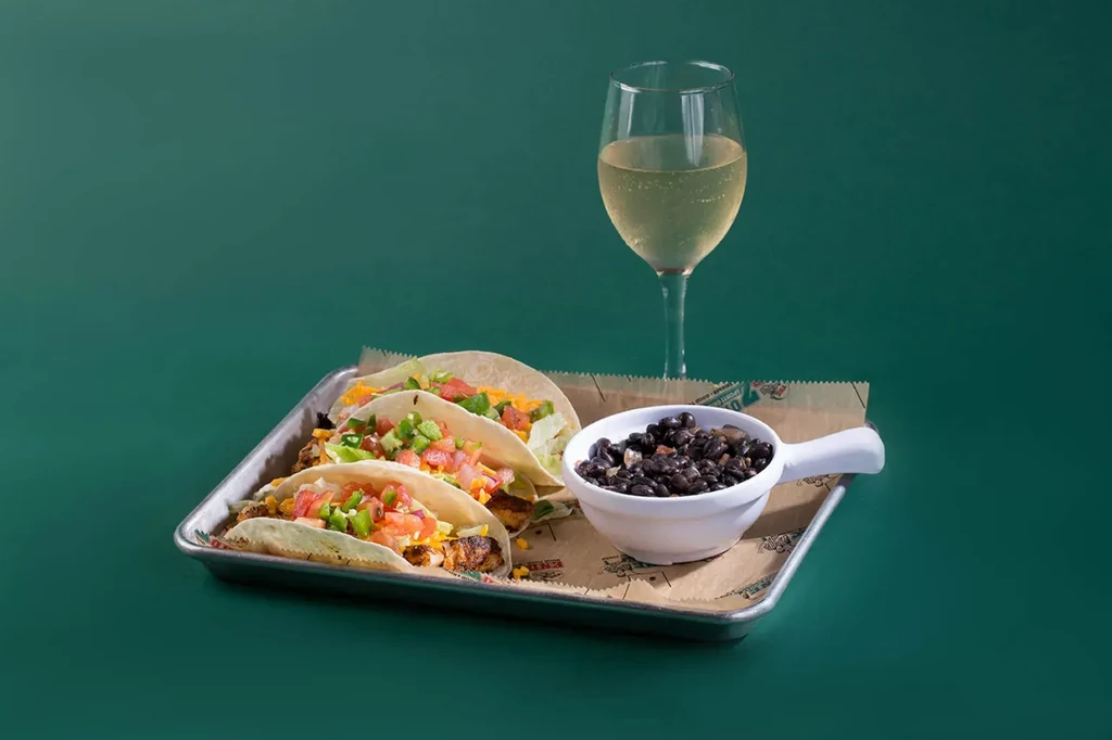 Three soft tacos, black beans, and white wine on a metal tray.