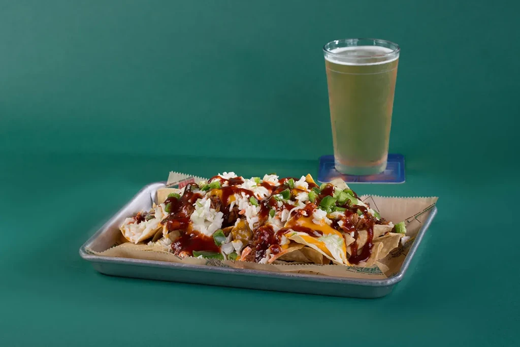 Loaded nachos with meat, slaw, peppers, and beer on a teal background.