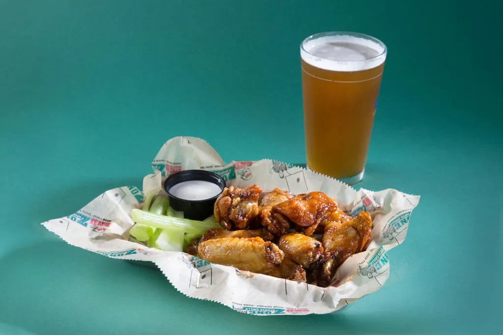 Basket of golden chicken wings, celery, dipping sauce, and an amber beer.