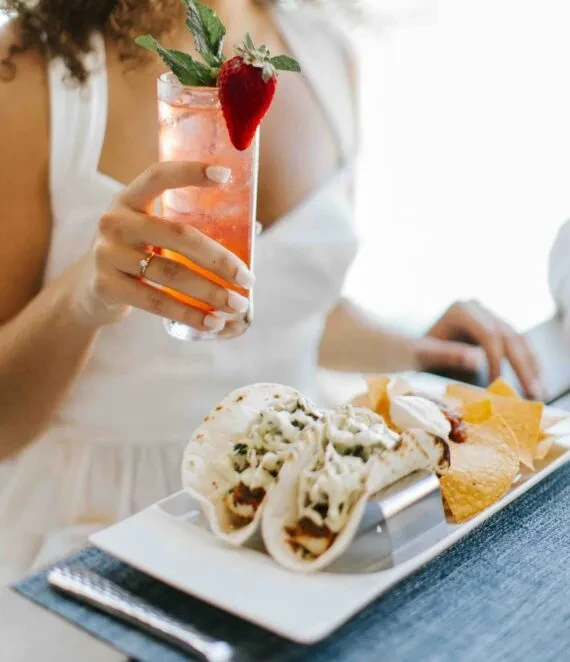 A hand holds a pink cocktail next to tacos, chips, and dip.