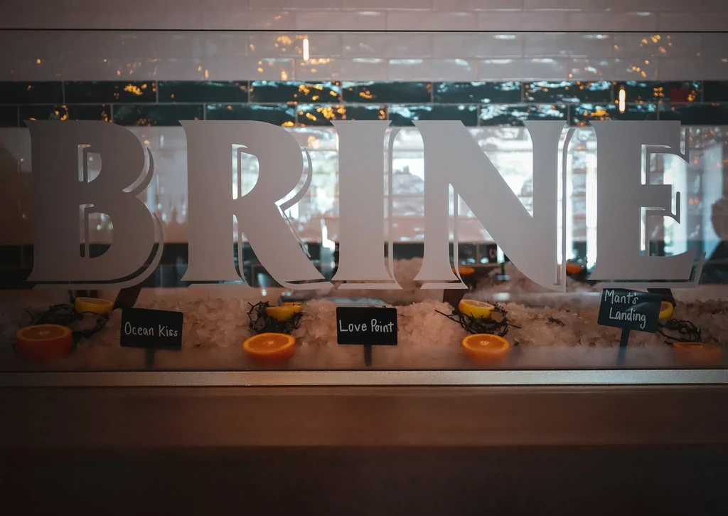 Fresh oysters on ice with citrus, 'BRINE' text, and variety signs.