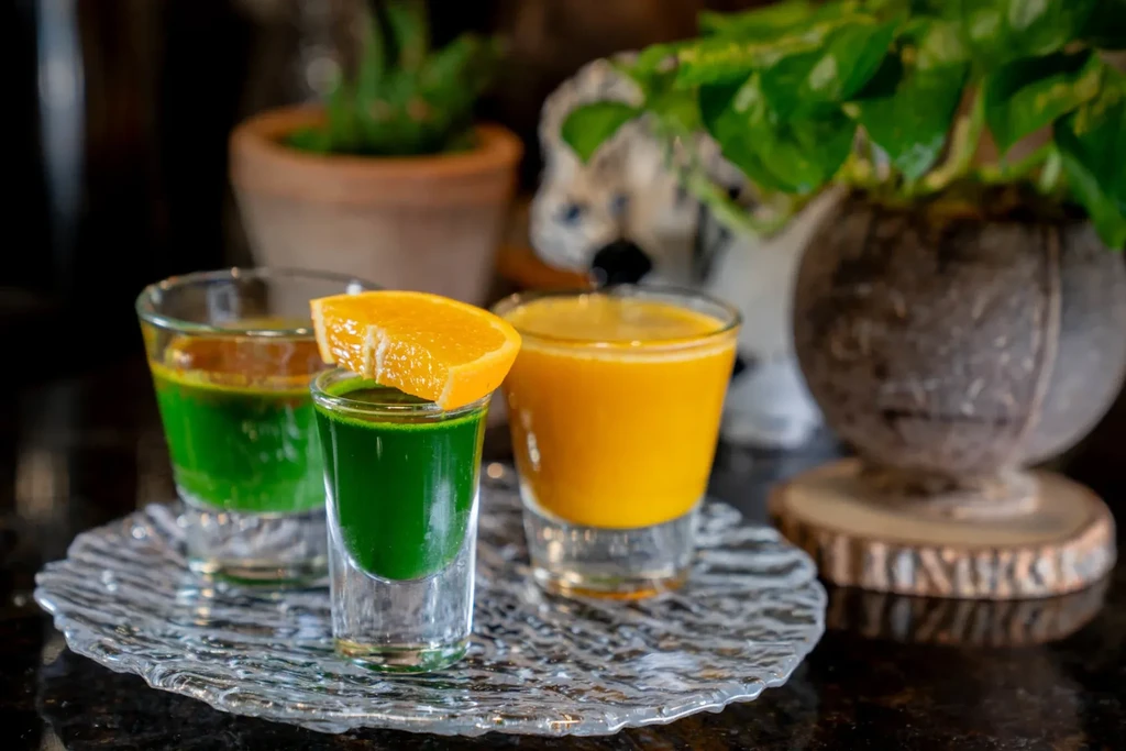 Three clear glasses with green and orange drinks on a textured glass plate.