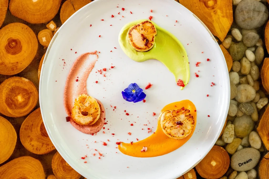 Three seared scallops, colorful purees, blue flower, wood, and river stones.