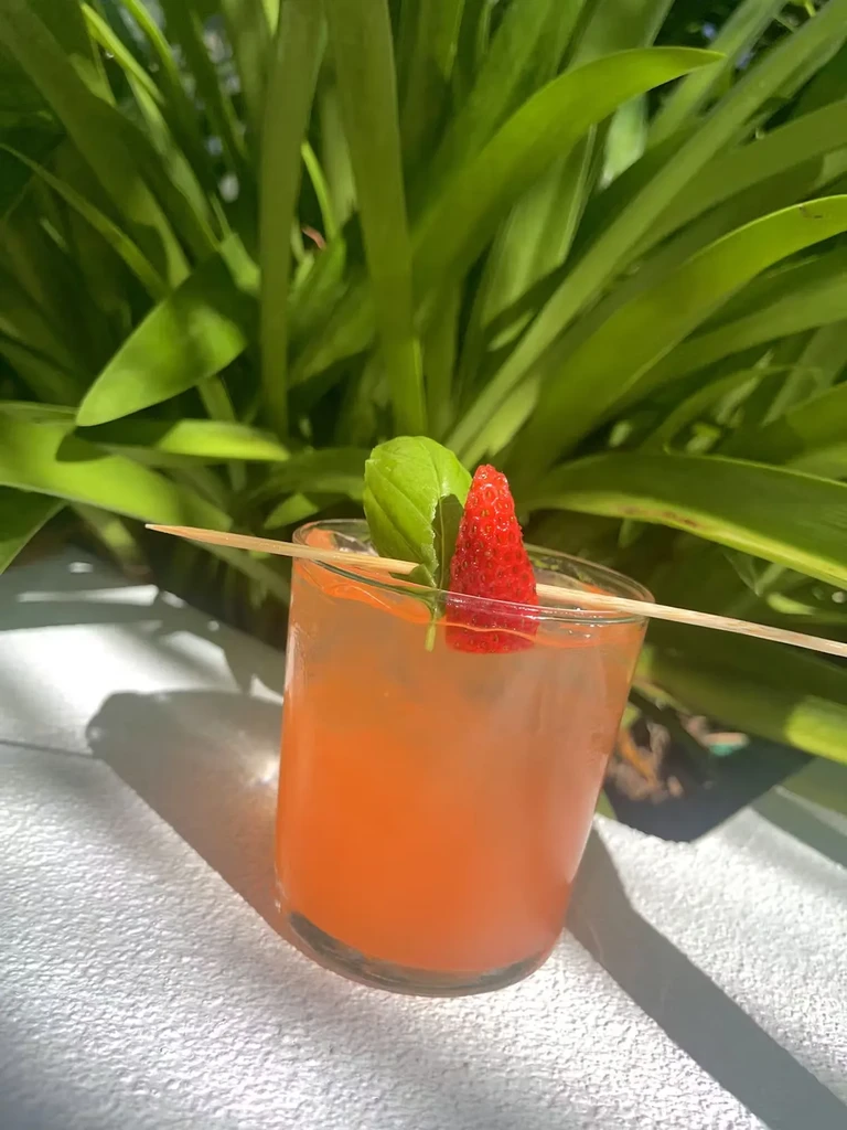 A vibrant reddish-orange cocktail garnished with basil and strawberry, outdoors in sunlight.