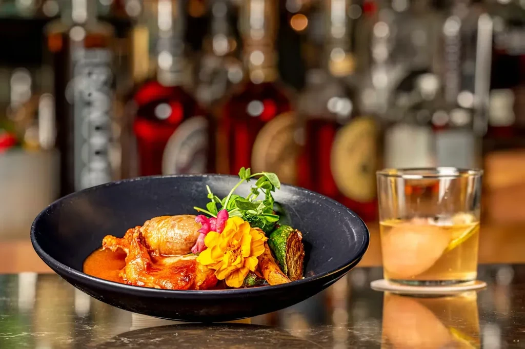 Plated dish with protein, colorful garnish, and amber cocktail with ice.