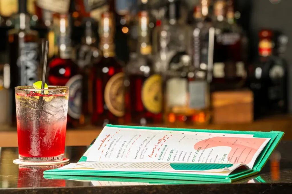 Red cocktail with lime, and open teal menu on bar counter.