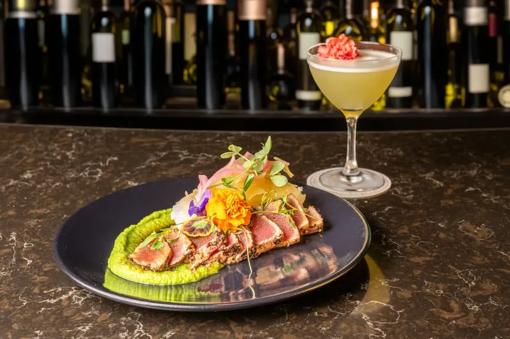 Plated seared tuna and yellow cocktail on a dark bar.