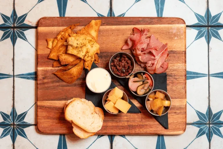 Overhead charcuterie board: chips, bread, dips, cheese, meat, pickled vegetables.