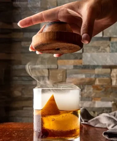 Amber drink with smoke rising, hand lifts smoking cap.