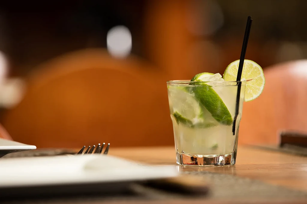 Clear lime cocktail, ice, lime wedges, straw on wooden table.