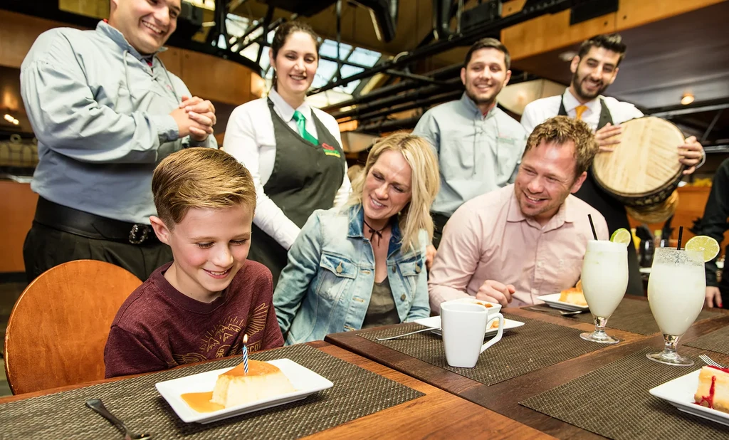 Smiling boy celebrates birthday with family, candlelit flan, and staff.