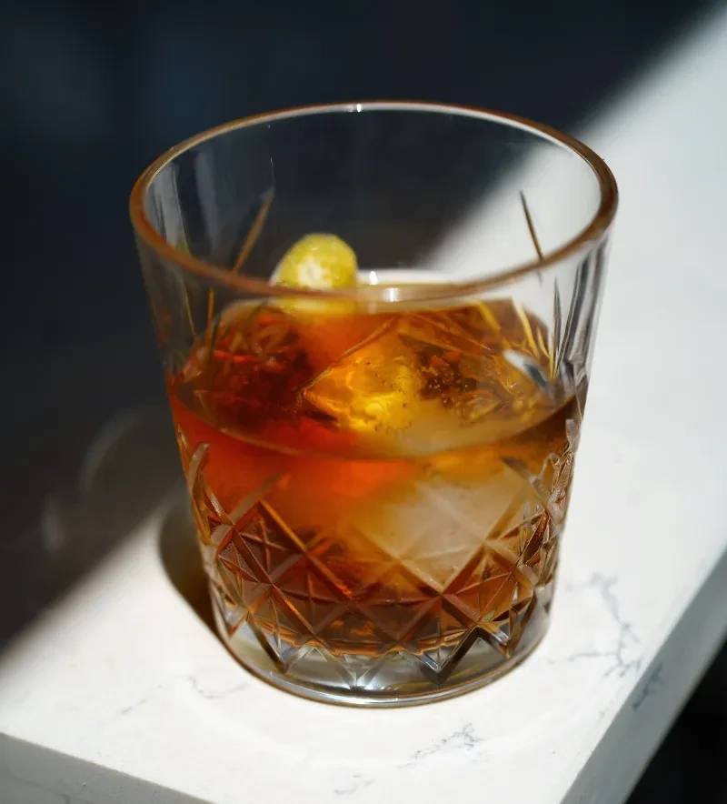 Dark amber cocktail with ice and lemon peel in a faceted glass.