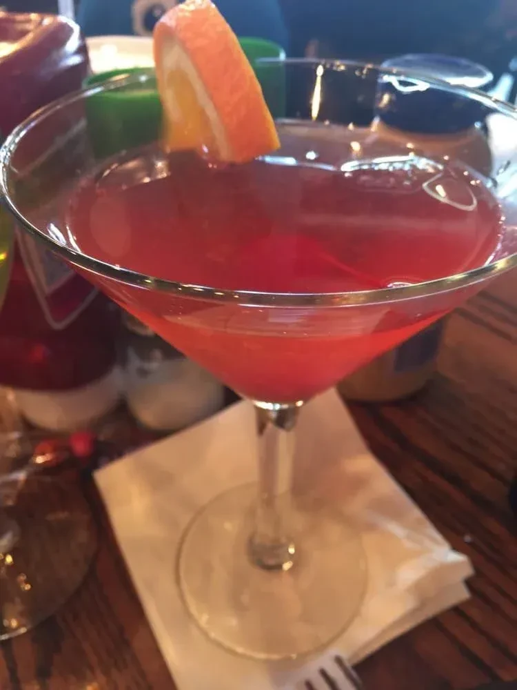 A vibrant red-pink cocktail in a martini glass with orange garnish.