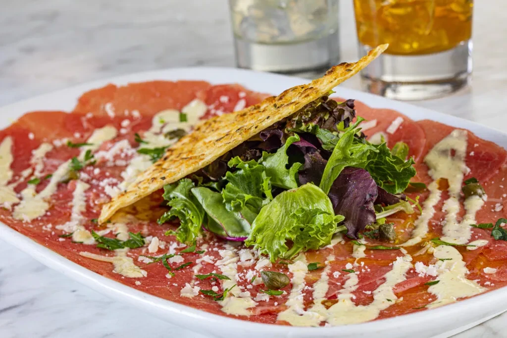 Artfully plated carpaccio with parmesan, capers, herbs, salad, and cracker.