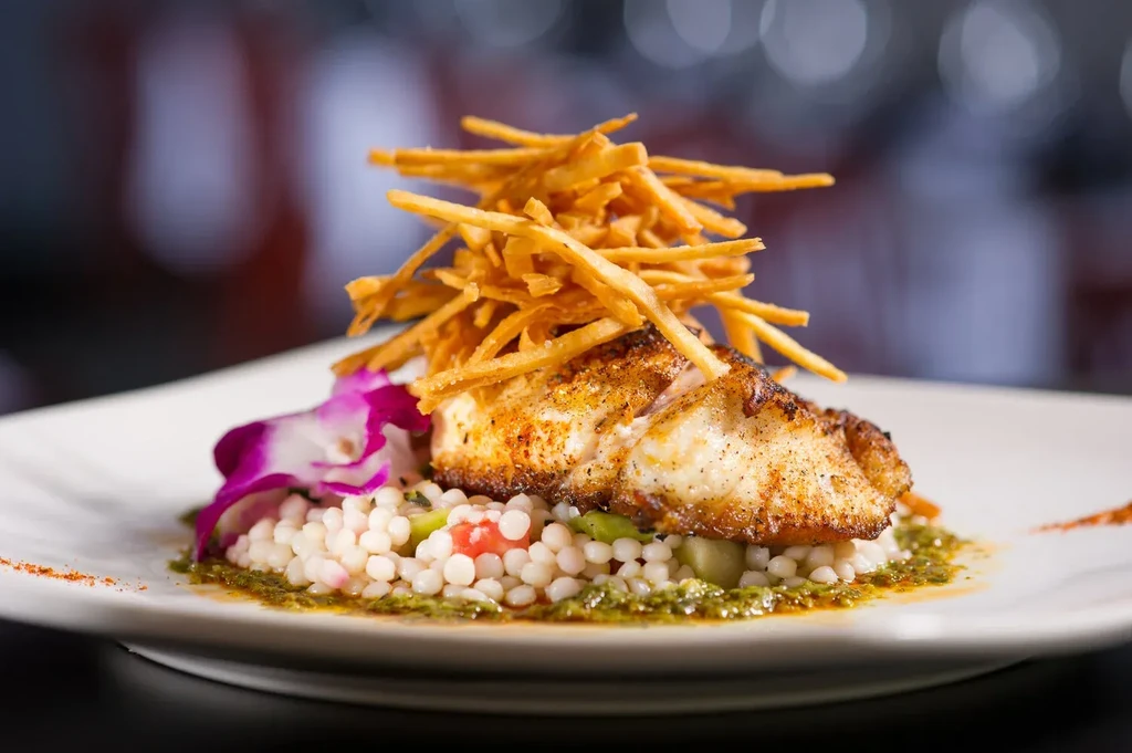 Plated seared fish, couscous, potato sticks, green sauce, and orchid garnish.
