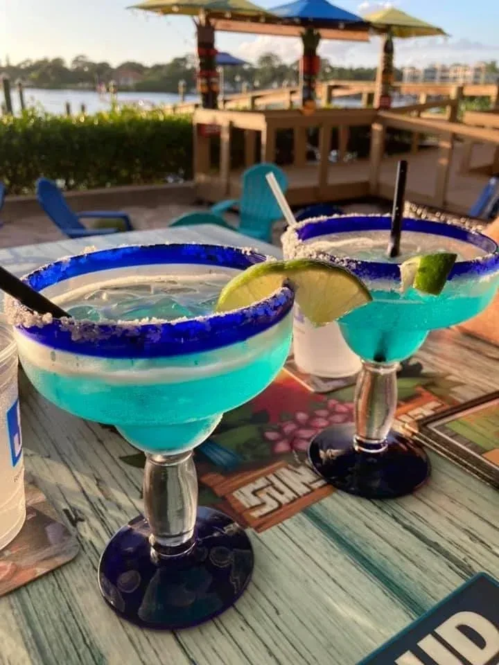Two vibrant turquoise margaritas on a table at an outdoor waterfront bar.