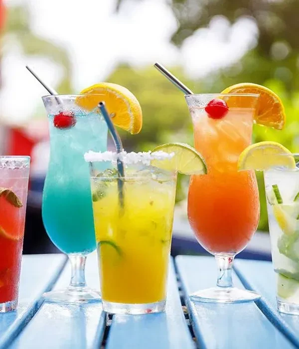 Five colorful garnished cocktails on a light blue wooden outdoor table.