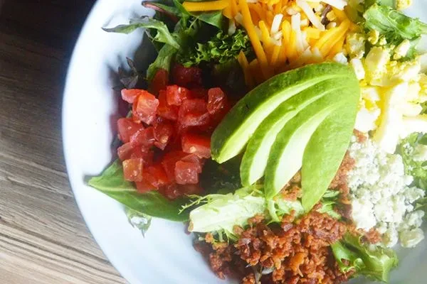 Cobb salad in white bowl with avocado, bacon, egg, and cheese.