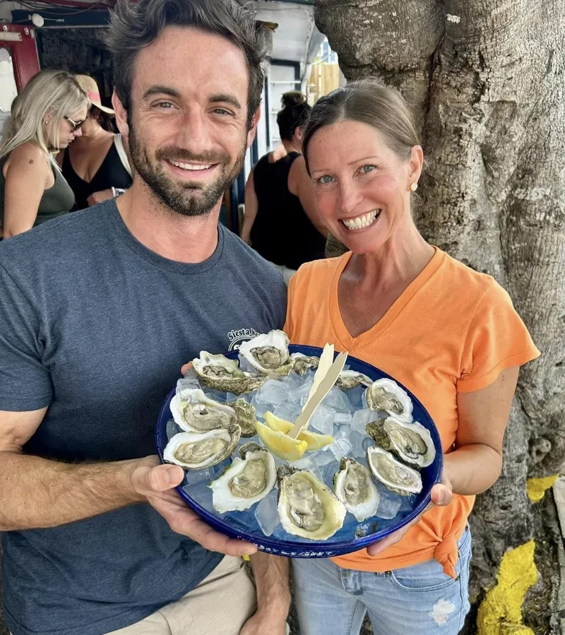 Smiling man and woman hold platter of raw oysters on ice.