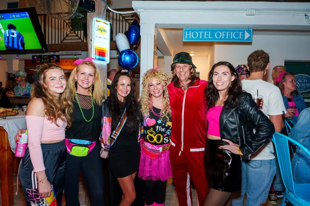 Six people in vibrant 80s attire smiling in a bar.