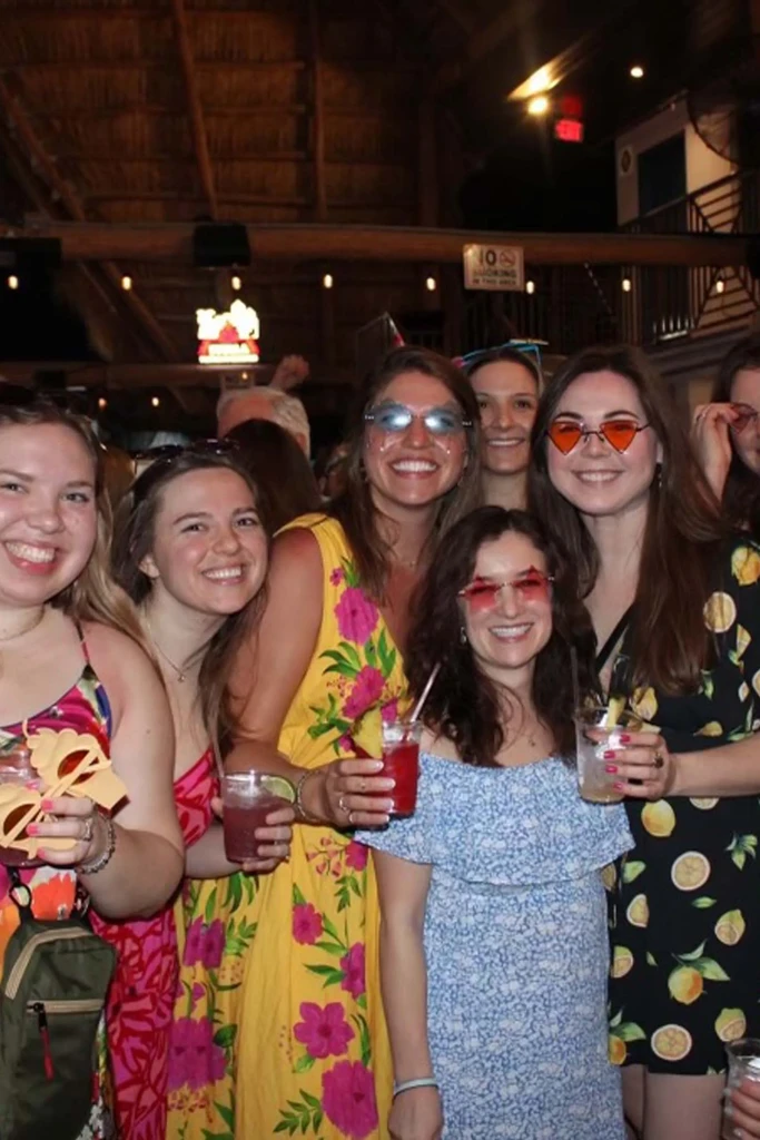 Six women smile, holding cocktails and wearing novelty sunglasses.