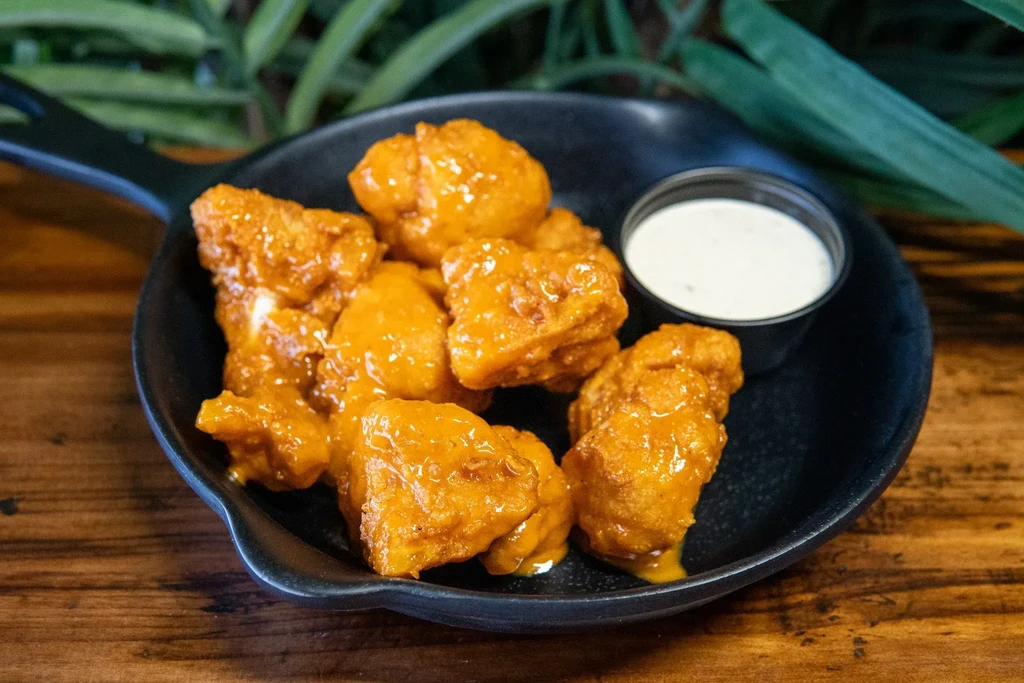Close-up: Golden chicken/cauliflower bites, white sauce, black pan on wood.