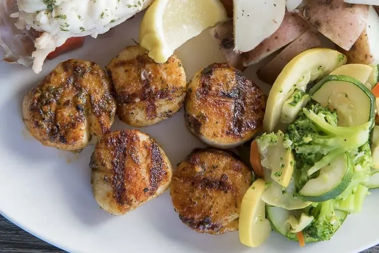 Grilled scallops, lobster, potatoes, and mixed vegetables on a white plate.
