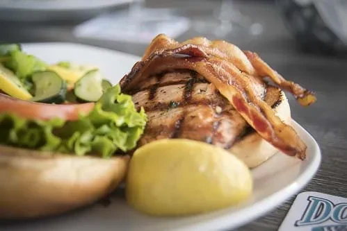 Angled close-up of a grilled sandwich with bacon, lettuce, tomato, and lemon.