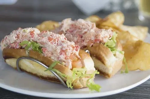 Two overflowing lobster rolls and golden potato chips on a white plate.