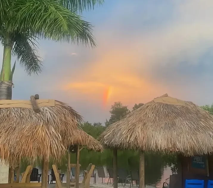 Tiki huts by calm water at dusk under a sunset rainbow.