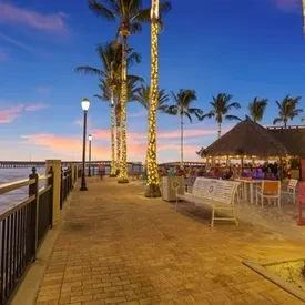 Tropical waterfront restaurant scene with string-lit palm trees at dusk.