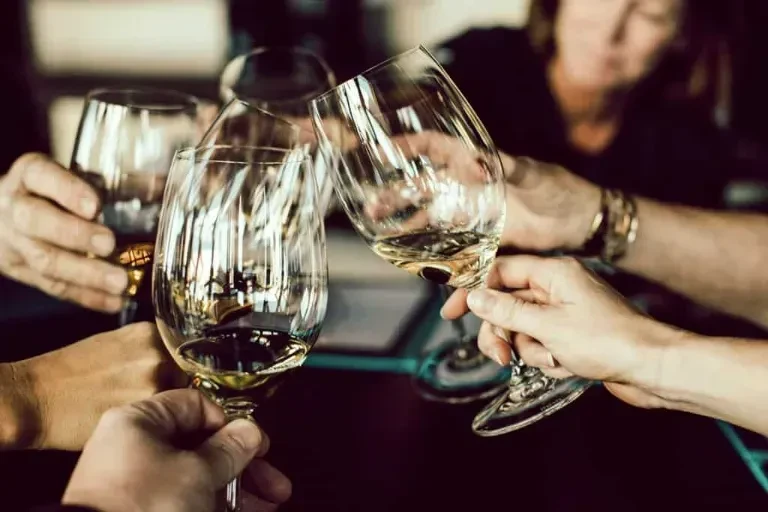 Four hands clinking stemmed wine glasses with light liquid.