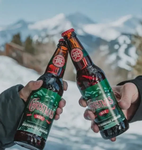Two hands clink Breckenridge Christmas Ale bottles in snowy mountains.