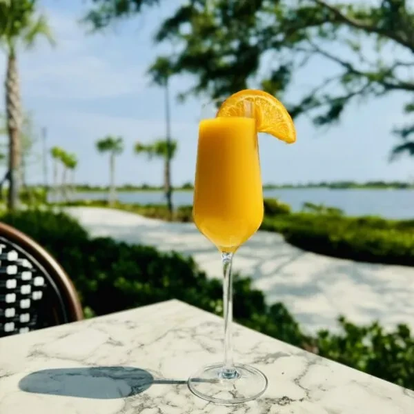 Orange drink in a flute with an orange slice by the water.
