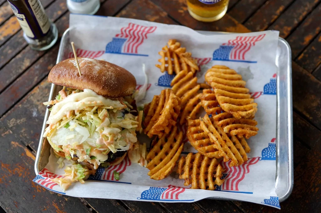Overhead: Burger with coleslaw, waffle fries, and patriotic tray on a wooden table.