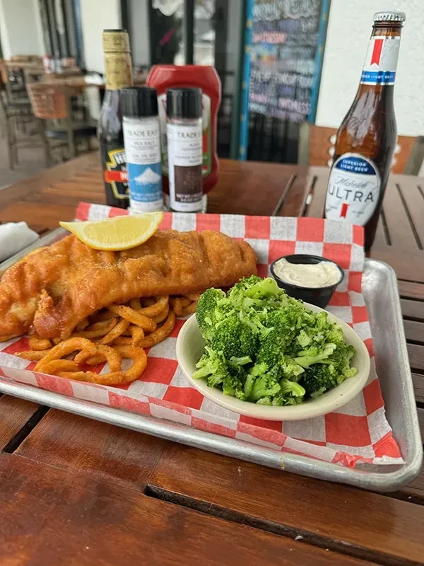 Golden battered fish, curly fries, broccoli, and beer on outdoor table.