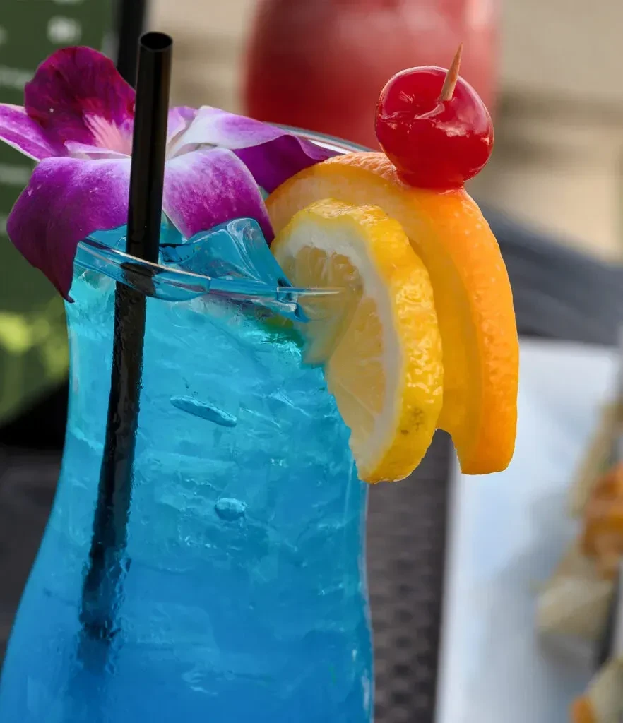 Vibrant blue cocktail with ice, orchid, lemon, orange, and cherry garnishes.