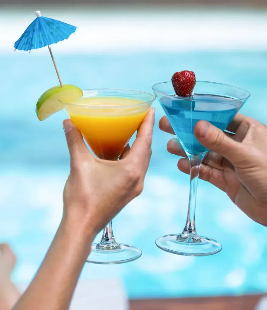Hands hold orange and blue cocktails in martini glasses at a pool.