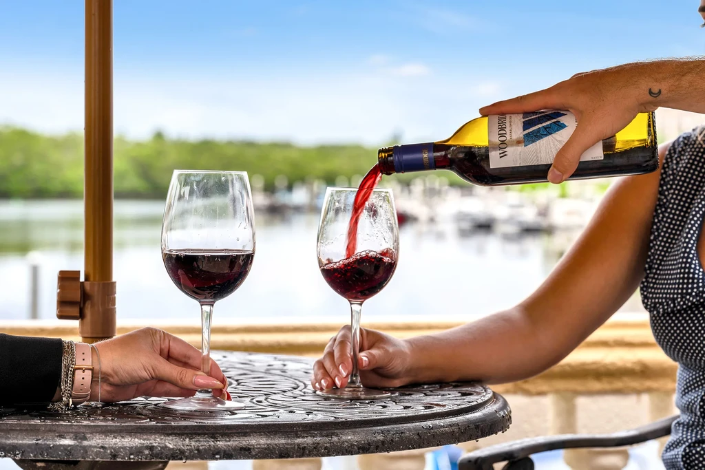 Hands pour red wine into a glass on a waterfront patio.