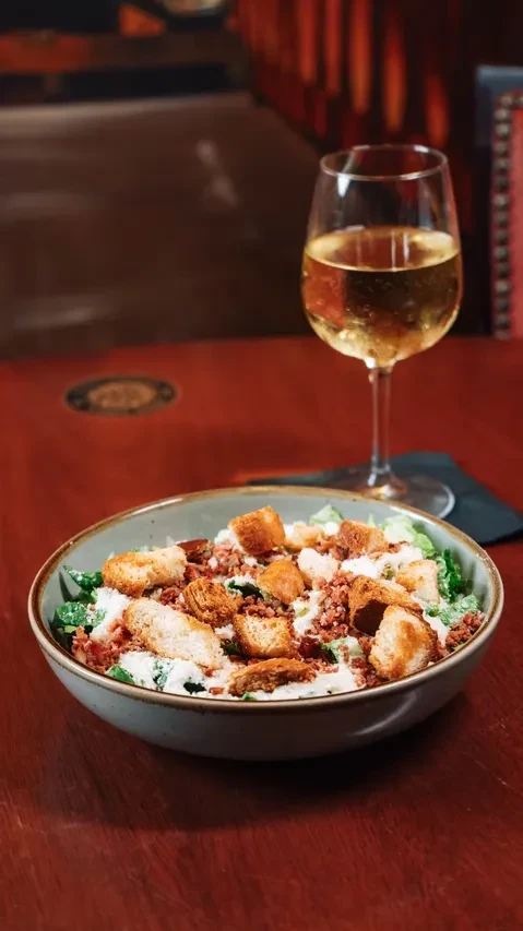 Close-up of green salad with croutons, bacon, cheese, and white wine.