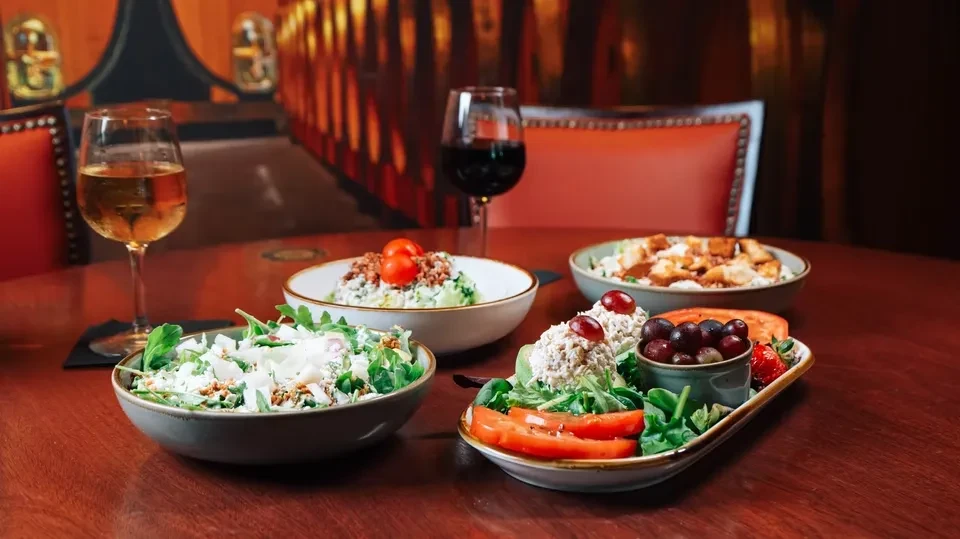 Four food dishes and two wines on a dark wood restaurant table.