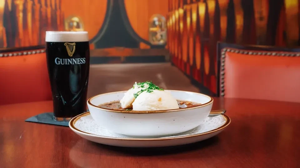 Dark stew with dumplings, herbs, and a Guinness stout on a table.