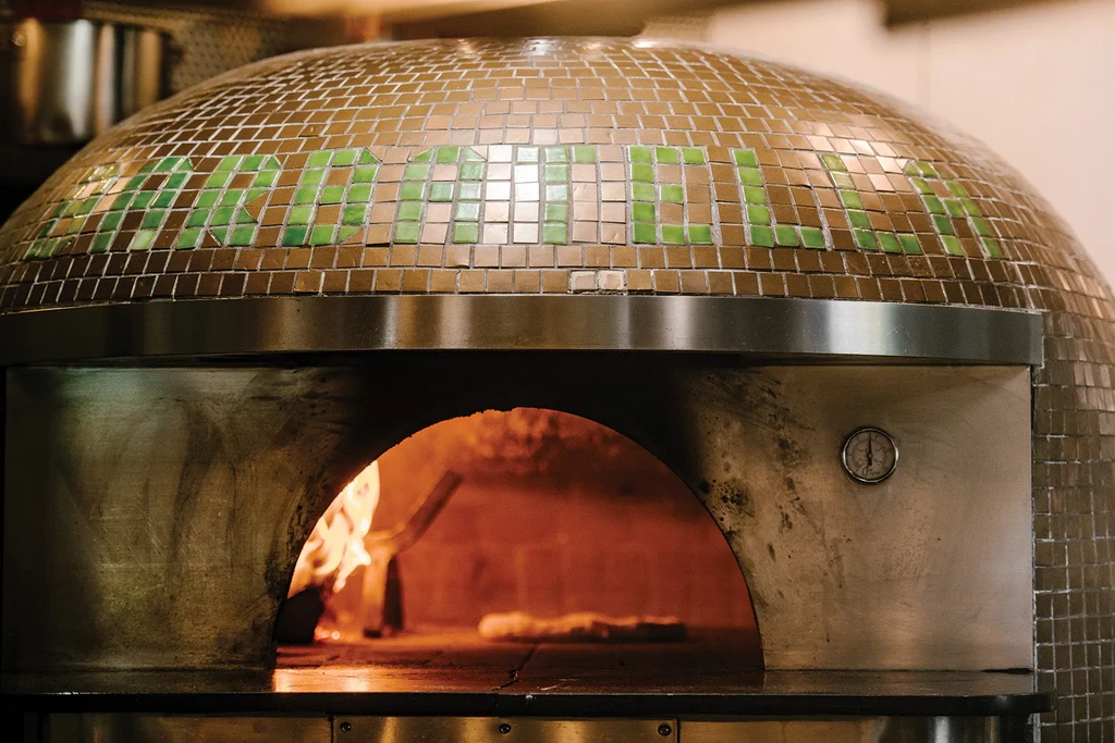 Dome-shaped mosaic pizza oven with internal flames baking a pizza.