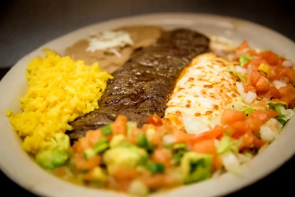 Meal with yellow rice, grilled steak, cheesy dish, beans, and toppings.