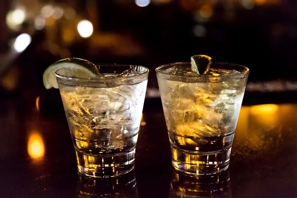 Two light drinks with ice and lime on a dark, reflective surface.