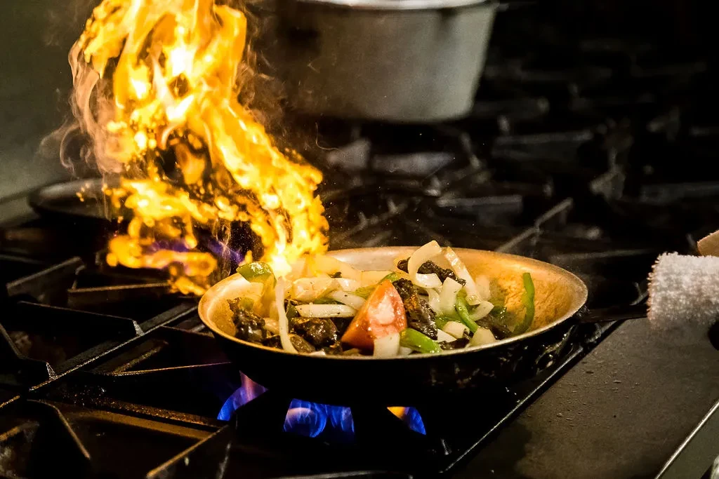 Meat and vegetables flambéing in a pan on a gas stove.