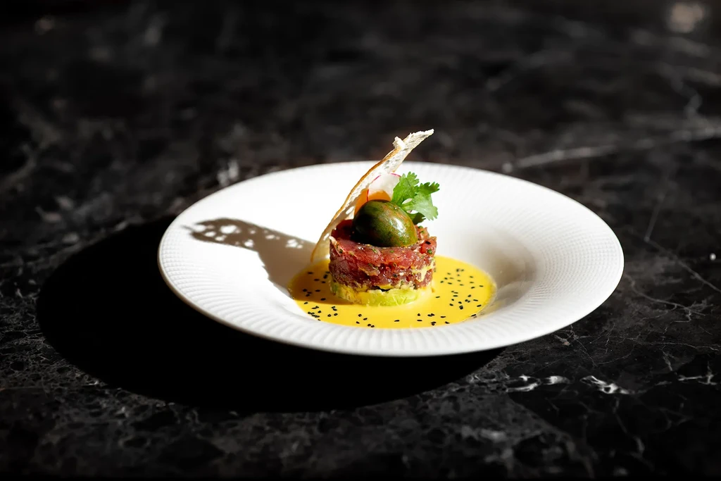 Cylindrical tuna tartare stack over avocado, with yellow sauce and garnish.