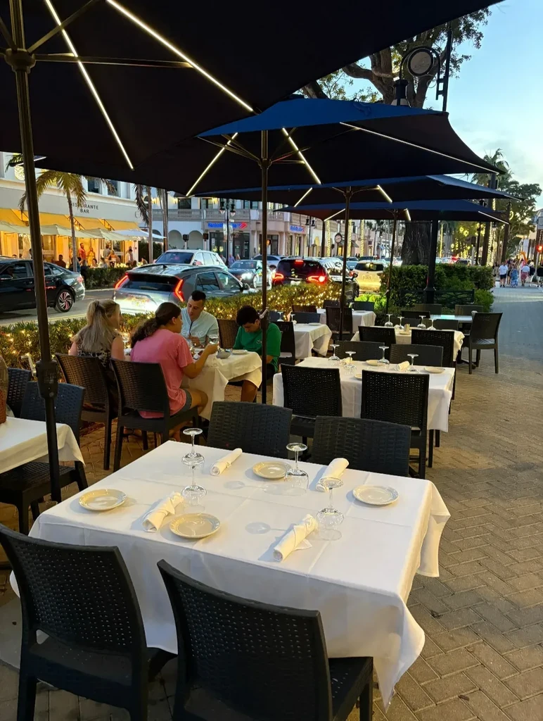 Outdoor restaurant dining area at dusk with diners and lighted umbrellas.