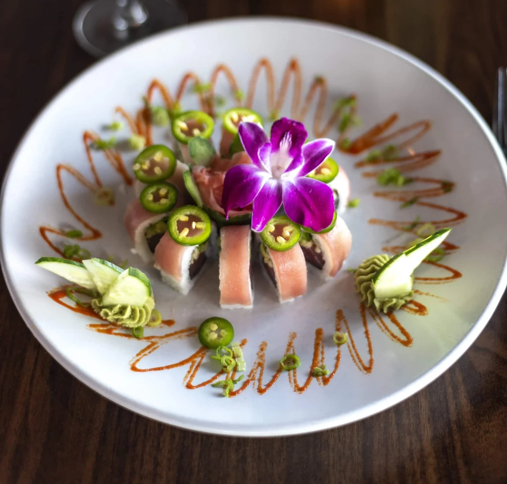 Plated sushi roll with jalapeños, purple orchid, and decorative sauces.